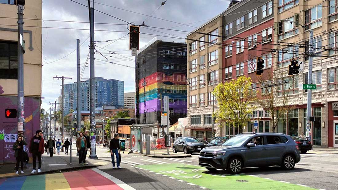 City street scene with pedestrians crossing a rainbow-painted crosswalk, cars waiting at a traffic light, and buildings in the background, including one with a large rainbow mural.
