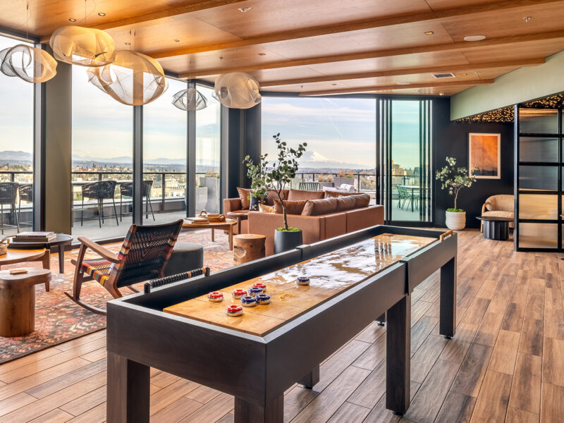 Modern lounge with a shuffleboard table, leather sofas, large windows, wooden floors, potted plants, and city views. Outdoor seating is visible through glass doors. Warm lighting and stylish decor create a cozy atmosphere.