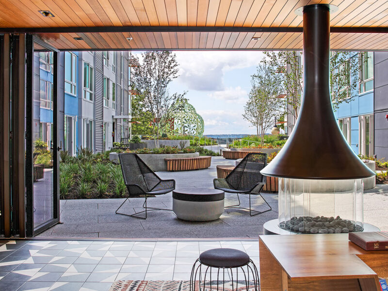 Modern outdoor patio with black chairs, a round table, wooden benches, greenery, and a decorative structure. Foreground shows a hanging fireplace and tiled floor inside. Sky and water visible in the distance.