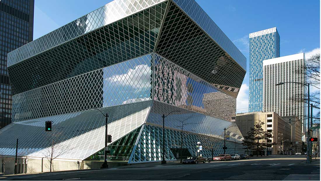 A modern glass building with geometric patterns stands on a city street, reflecting clouds and nearby skyscrapers under a blue sky.