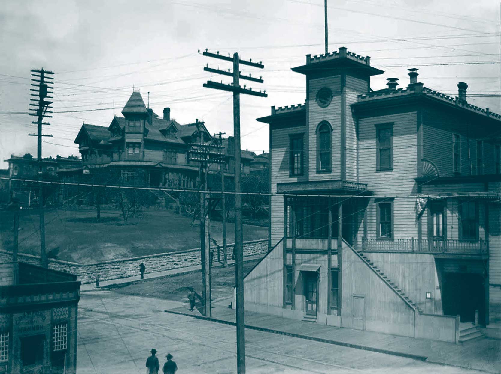 Historic black-and-white photo of a two-story wooden building at a street corner, with utility poles and wires, and a large Victorian mansion on a hill in the background.