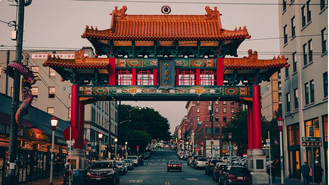 A vibrant, traditional Chinese gate stands over a city street lined with cars and buildings, with a decorative dragon on the left and people walking on the sidewalks.