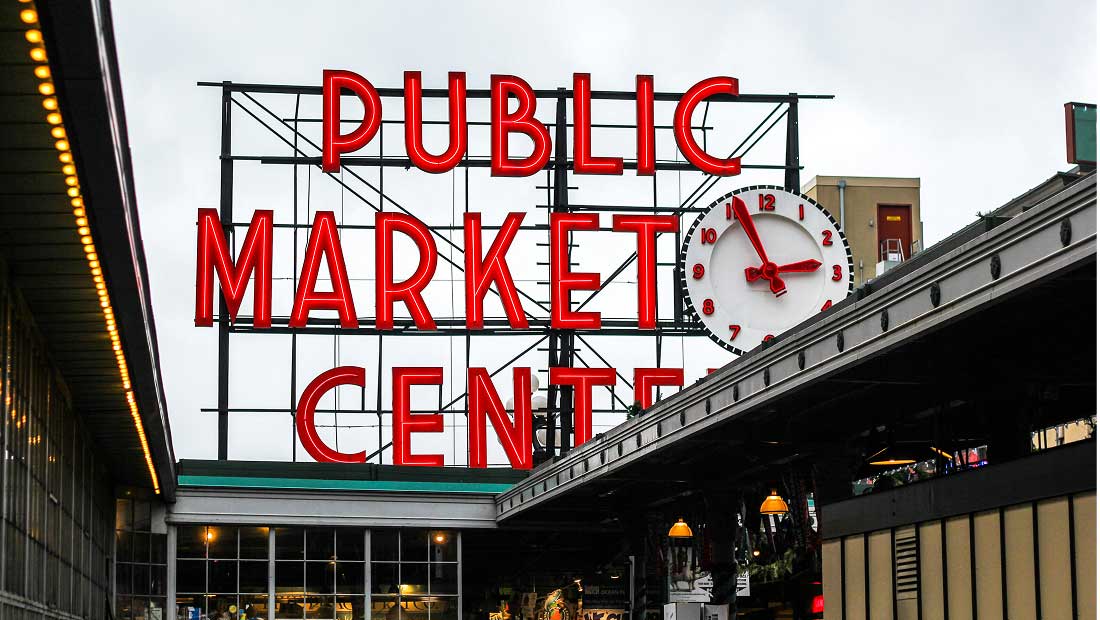 Large red neon Public Market Center sign with a white clock is displayed above a market entrance, seen on a cloudy day. The sign and clock are landmarks of Seattles Pike Place Market.
