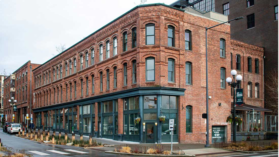 A large, historic red brick building with many windows sits on a street corner. The lower level has green-framed storefronts, and there are cars parked along the street. The sky is overcast.