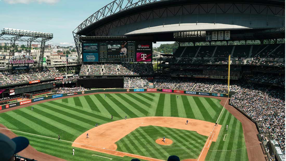 A baseball game underway in a large stadium with a partially open roof and a vibrant green field, filled with fans in the stands under sunny skies.
