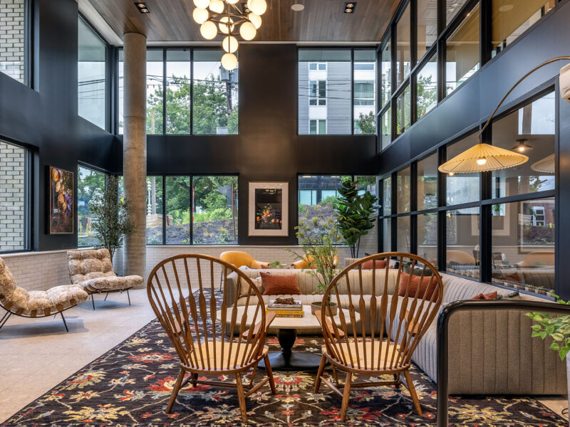 A modern lounge features large windows, wooden chairs, a gray sofa, a floral rug, plants, and contemporary lighting. The space is bright and open with stylish decor and natural light.